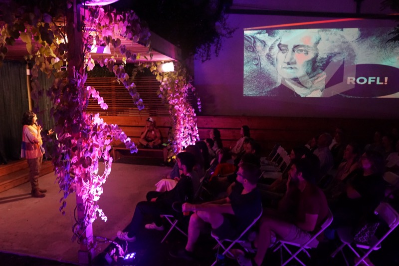 An outdoor film night in the garden, an audience seated in the dark beside vines lit up in pink.