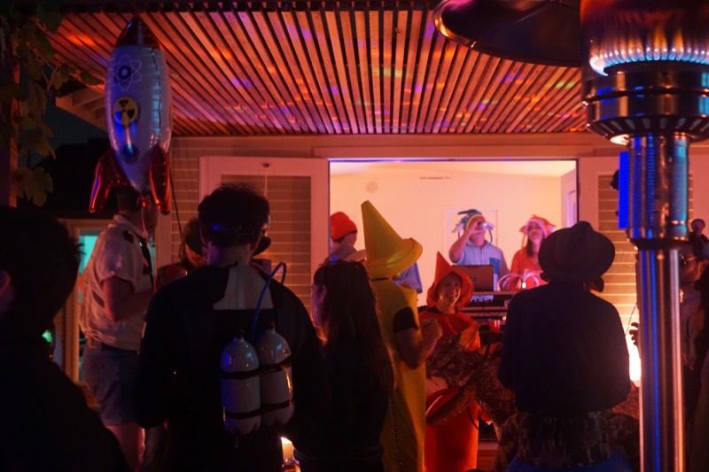 A costume party under the pergola at night, warm light and a patio heater among the guests.