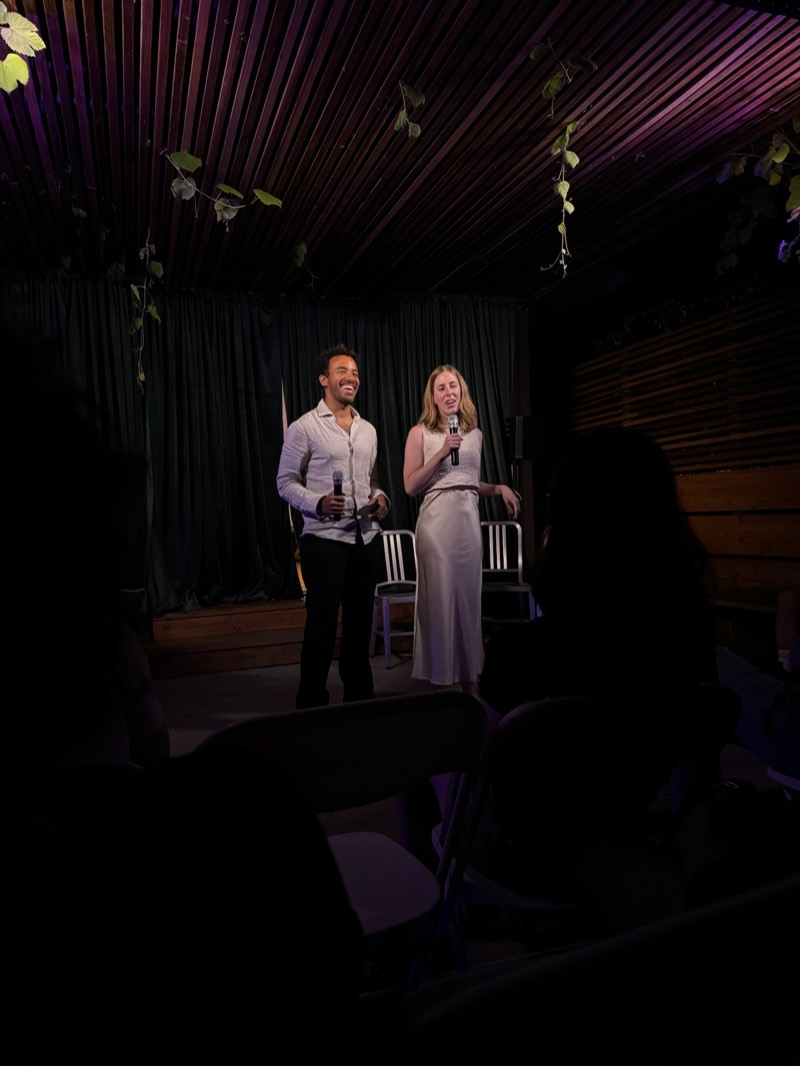 Phoebe and Brian on stage with microphones at a night event under the pergola.