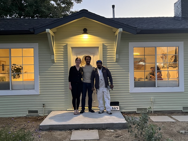 Three people standing arm in arm on the front step of the newly painted house at dusk.