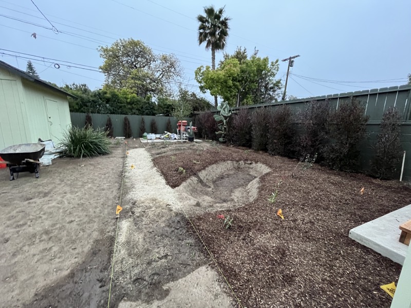 The back yard graded and mulched, with irrigation flags marking planting positions and a wheelbarrow standing by.