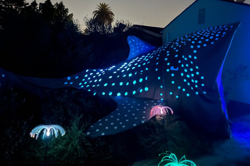 A large glowing manta ray sculpture covered in blue lights installed in the garden at night, with lit jellyfish below.