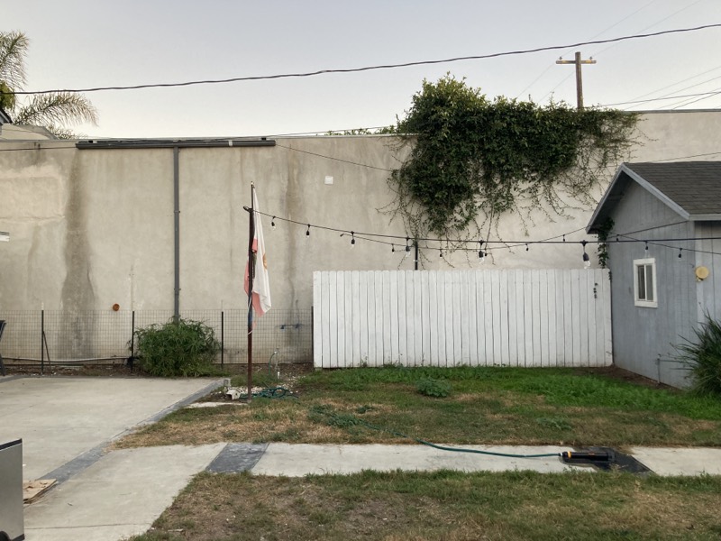 The back yard before the redesign: a bare concrete slab, patchy grass, a white fence, and a tall blank stucco wall.